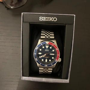 Seiko SKX009 - Säljer en Seiko dykarklocka med en robust stållänk och en boett i silver. Urtavlan är blå med tydliga markeringar och en röd-blå vridbar bezel. Klockan har dag- och datumvisning och är perfekt för den som gillar en sportig stil.