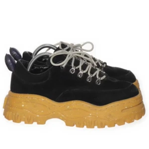 Svarta sneakers med tjock sula - Snygga svarta sneakers med en rejäl, gul sula och snörning. Perfekta för en cool och trendig look. Skorna har en unik design med metallöglor för snörena.