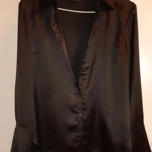 Svart satinblus - Elegant svart skjorta  i satin med långa ärmar och klassisk krage. Blusen har en stilren design med knappar framtill och vid ärmsluten. 
