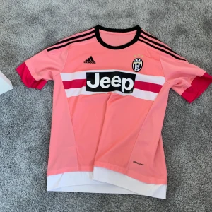 Rosa Juventus fotbollströja från Adidas - Snygg rosa fotbollströja från Adidas med svart och vit Jeep-logga på bröstet. Tröjan har korta ärmar med svarta ränder på axlarna och en vit och röd detalj över bröstet. Perfekt för fotbollsfans som vill sticka ut på planen!