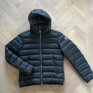 Svart dunjacka från Colmar - Snygg svart dunjacka från Colmar med huva och dragkedja. Jackan har ett quiltat mönster och är perfekt för kyligare dagar. Den har en stilren design med en logotyp på ärmen.