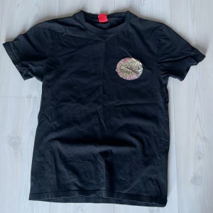Svart t-shirt från Santa Cruz - Säljer en svart t-shirt från Santa Cruz med ett färgglatt tryck på bröstet och ryggen. T-shirten är kortärmad och tillverkad i 100% bomull. Perfekt för en avslappnad stil.