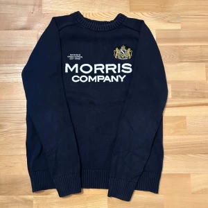 Svart tröja från Morris Company - Säljer en svart tröja från Morris Company med broderad logotyp och text på framsidan. Tröjan är tillverkad i 100% bomull och har långa ärmar samt ribbade muddar vid ärmslut och nederkant.