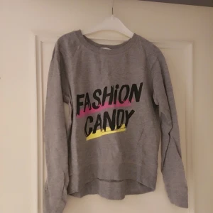 Grå  tröja till flickor med tryck från H&M - Säljer en grå tröja från H&M med texten 'Fashion Candy' i svart, rosa och gul. Tröjan har långa ärmar och en rund halsringning. Perfekt för en avslappnad stil.Storlek är 140,10 år