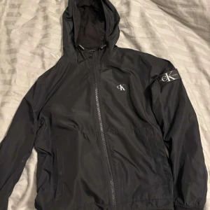 Svart vindjacka från Calvin Klein Jeans - Snygg svart vindjacka från Calvin Klein Jeans med huva och dragkedja. Jackan har logotypdetaljer på bröstet och ärmen. Perfekt för blåsiga dagar och ger en stilren look.