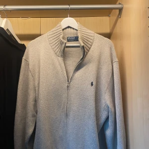 Grå stickad tröja från Polo Ralph Lauren - Säljer en stilren grå stickad tröja från Polo Ralph Lauren med dragkedja och ribbad krage. Tröjan har långa ärmar och en liten broderad logotyp på bröstet. Den är i bra skick