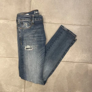Blå jeans från Jack & Jones - Snygga blå jeans från Jack & Jones med slitna detaljer och en klassisk femficksdesign. De har en knappgylf och är perfekta för en avslappnad stil. Passformen är slim och de är tillverkade i ett bekvämt denimtyg.