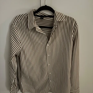 Skjorta - Snygg brun och vit randig skjorta. Skjortan har långa ärmar och knappar framtill. Perfekt för en stilren look.