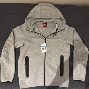 Nike Tech Fleece Hoodie Zip Up - Jag säljer en grå Nike tech fleece hoodie zip up. Den är helt ny med prislapp och är inte använd. Jag har bara provat en gång i några minuter. Den är i storlek M. Skriv till mig om du har några frågor!