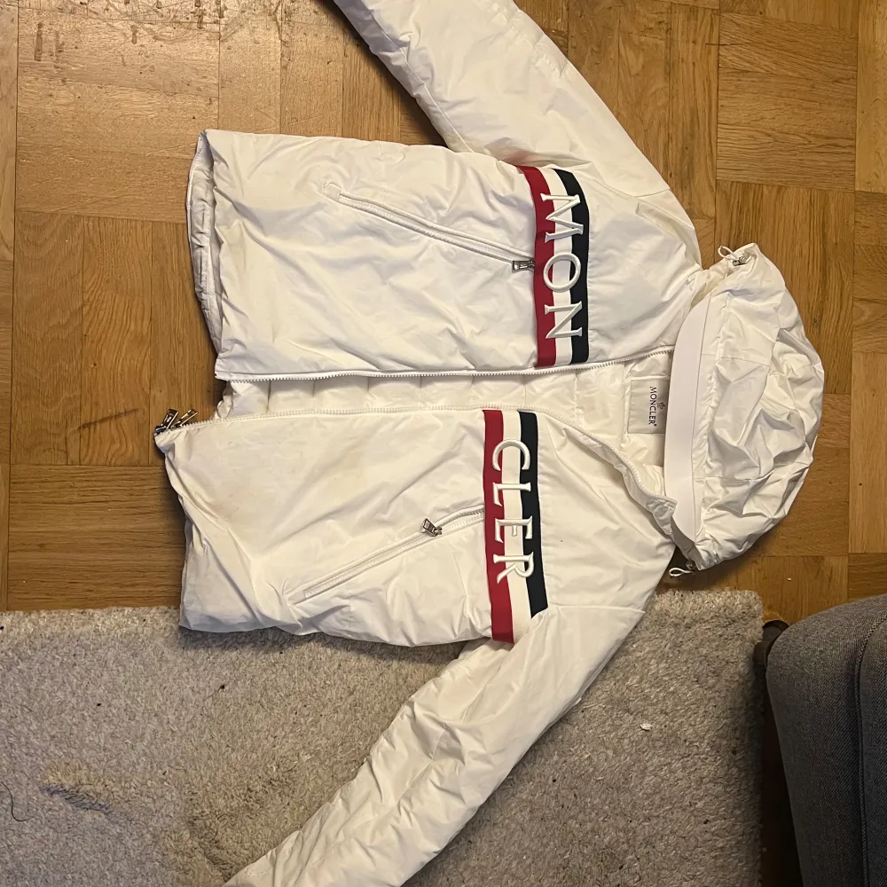 Säljer en snygg vit pufferjacka från Moncler med huva och dragkedja. Jackan har Moncler-logga på bröstet och ett stilrent band i rött och blått. Perfekt för kyliga dagar!. Takit.