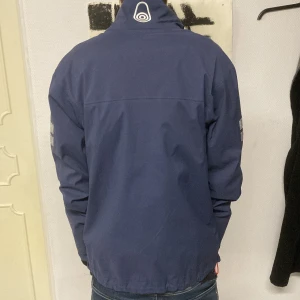 Blå jacka från Sail Racing - Säljer en blå jacka från Sail Racing med dragkedja och logotypdetaljer på bröstet och ärmen. Jackan har en hög krage och är långärmad, perfekt för kyligare dagar. Den har en stilren design som passar bra till jeans.