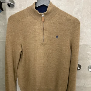 Beige/ljusbrun ulltröja från Morris - Säljer en stilren beige tröja från Morris i 100% merinoull. Tröjan har en halv dragkedja och en liten broderad detalj på bröstet. Perfekt för kyligare dagar med sin mjuka och varma känsla.