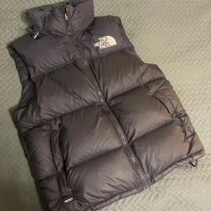 Svart dunväst från The North Face - Säljer en svart dunväst från The North Face med dragkedja och hög krage. Västen har märkets logga på bröstet och ryggen. Perfekt för kyligare dagar.
