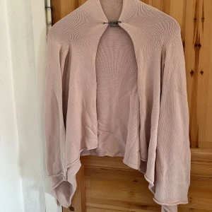 Ljusrosa kofta från Hunkydory - Säljer en elegant ljusrosa kofta från Hunkydory Essentials. Den är stickad och har långa ärmar med en unik säkerhetsnål som man kan använda för att knäppa koftan.. Perfekt för att hålla sig varm och stilren under kyligare dagar.