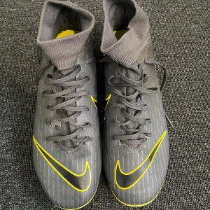Grå och gula fotbollsskor från Nike - Säljer ett par grå fotbollsskor från Nike med gula detaljer . Skorna har en strumpliknande krage för extra stöd och en slitstark sula med dobbar för bra grepp på planen. Perfekta för fotbollsträning och matcher.