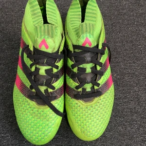 Gröna fotbollsskor från Adidas - Snygga gröna fotbollsskor från Adidas med rosa och svarta detaljer. Skorna har snörning och en mönstrad yta för bättre grepp på planen. Perfekta för fotbollsspelare som vill sticka ut.