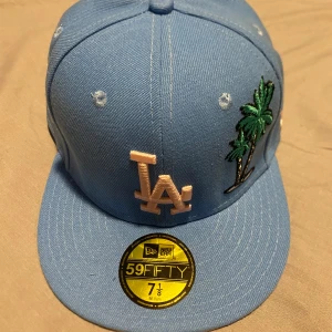New Era keps  -  blå keps från New Era med broderad LA-logga och palmer på framsidan. Kepsen har även ett broderat emblem. Den är i storlek 7-1/8 och har en rak skärm. Helt Ny. Pris kan diskuteras. Storleken i cm är 57cm.