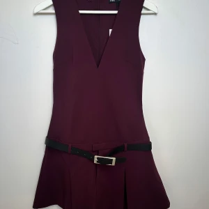 Vinröd klänning med bälte från Zara - Snygg vinröd klänning från Zara med v-ringning och ett stilrent svart bälte i midjan. Klänningen är ärmlös och har en kort längd som ger en elegant look. Perfekt för att sticka ut med stil! Prislapp kvar