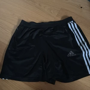 Svarta träningsshorts från Adidas - Snygga svarta träningsshorts från Adidas med de klassiska tre vita ränderna längs sidorna. Shortsen har en elastisk midja och dragkedjefickor för säker förvaring. Perfekta för sport och träning.