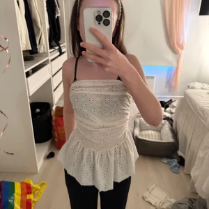 Vit axelbandslös topp - Säljer en söt vit axelbandslös topp med broderade detaljer och volangkant. Perfekt för sommarens varma dagar. Toppen har ett fint hålmönster som ger en romantisk känsla.