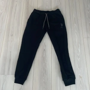 Svarta mjukisbyxor från USPA Sport - Snygga svarta mjukisbyxor från USPA Sport med broderad logga på benet. Byxorna har en bekväm passform med elastisk midja och snörning. Sköna och bekväma mjukisbyxor perfekta för en avslappnad stil. 