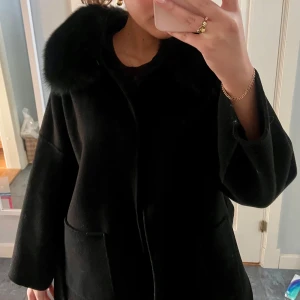 Svart kappa med päls luva  - Svart kappa med en pälskrage och knytbälte i midjan. Perfekt för kyliga dagar under höst o vinter. Använd fåtal gånger. Passar både M och S.