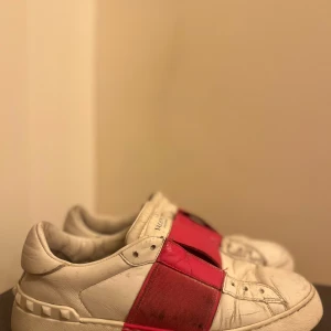 Valentino Garavani sneakers med röd detalj - Snygga Valentino Garavani sneakers i vitt skinn med en iögonfallande röd detalj över ovansidan. Skorna har en klassisk design med rund tå och subtila nitar på hälen för en extra touch av stil.