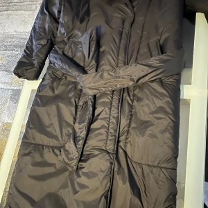 Svart dunjacka med bälte S i storlek  - Säljer en svart lång dunjacka med bälte i midjan. Jackan har långa ärmar och en stilren design som passar perfekt för kyliga dagar. Den är både praktisk och snygg, redo att hålla dig varm hela vintern.