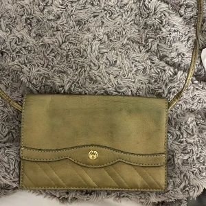 Olivgrön axelväska med gulddetalj - Snygg olivgrön axelväska i skinn med quiltad design och en elegant guldfärgad knappdetalj. Perfekt för att ge en touch av stil till din outfit. Väskan har en smidig axelrem och en praktisk storlek för dina nödvändigheter.