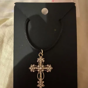 Halsband med korshänge - Egengjort halsband med ett korshänge dekorerat med pärlor. Hänget har en detaljerad design och hänger på en svart snodd. Perfekt för att ge en stilfull touch till din outfit.