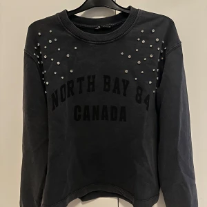 Svart sweatshirt med pärlor - Snygg svart sweatshirt med texten 'North Bay 84 Canada' tryckt på framsidan. Tröjan har långa ärmar och är dekorerad med pärlor över axlarna och bröstet. Perfekt för en avslappnad stil med en touch av elegans.