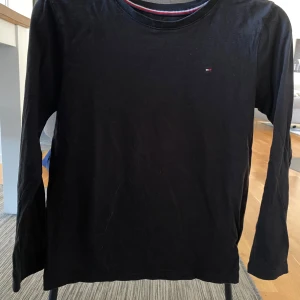 Svart långärmad tröja från Tommy Hilfiger - Säljer en stilren svart långärmad tröja från Tommy Hilfiger. Tröjan har en liten logotyp på bröstet och en randig detalj i kragen i rött, vitt och blått. Perfekt för en avslappnad och snygg look.