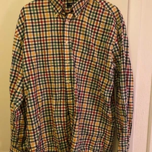 Rutig skjorta från GANT - Säljer en färgglad rutig skjorta från GANT i storlek 2XL. Skjortan har en klassisk krage och knappar framtill. Perfekt för en avslappnad stil med sin casual fit. Den har långa ärmar och en bröstficka.