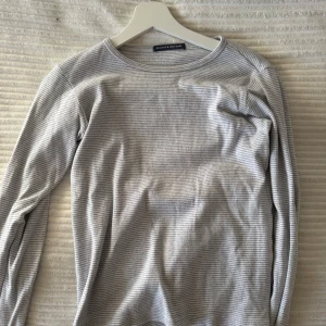 Grå randig långärmad tröja från brandy Melville - Säljer en stilren grå randig långärmad tröja från brandy Melville. Perfekt för kyligare dagar med sin mjuka och bekväma passform. Tröjan har en rund halsringning och är tillverkad i ett lätt material.