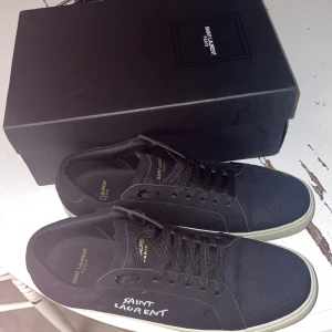 Svarta sneakers från Saint Laurent - Snygga svarta sneakers från Saint Laurent med vit text på sidan. Skorna har en klassisk design med snörning och en vit sula som ger en stilren look. Perfekta för en trendig vardagsstil.