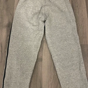 Grå mjukisbyxor från Adidas - Säljer ett par grå mjukisbyxor från Adidas med svarta ränder längs sidorna och en liten logga på framsidan. Byxorna har en bekväm passform med resår och snörning i midjan samt muddar vid bensluten.