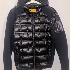 Svart pufferjacka med huva - Säljer en snygg svart pufferjacka från Parajumpers. Den har en quiltad design med dragkedja och en cool patch på ärmen. Jackan är perfekt för höst och vinter, och har en skön huva för extra värme. Den är i nyskick och redo för nya äventyr! Köpt iår för 6500kr