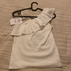 Vit oneshoulder topp från WE, PRIS DISKUTERAS !!!❤️ - Säljer en superfin vit oneshoulder topp från WE i ekologisk bomull. Toppen har en snygg volangdetalj längs axeln som ger en extra touch. Perfekt för sommardagar eller festliga tillfällen! 🌟