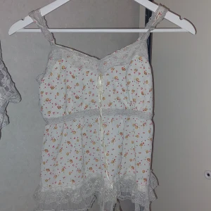 Blommig linne med spetsdetaljer - Söt och romantisk linne i vitt med små blommor i rosa och rött. Toppen har spetsdetaljer vid halsringningen och nederkanten, samt en liten rosett framtill. Perfekt för varma sommardagar eller som ett sött lager under en kofta.