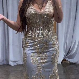Grå/Guld Aftonklänning - En vacker klänning med guld spets och en figurformad silhuett. Klänningen har en djup V-ringning och en spetsdetalj i ryggen. Den är perfekt för en speciell tillställning såsom bröllop eller även nyår