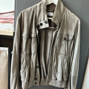 Beige mockajacka från Brunello Cucinelli - Säljer en snygg beige mockajacka från Brunello Cucinelli. Jackan har en stilren design med knappar framtill och flera praktiska fickor. Den är tillverkad i Italien och har en mjuk och lyxig känsla. Perfekt för höst och vår!