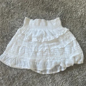 Vit kjol från Neo Noir - Säljer en superfin vit kjol från Neo Noir. Den har fina volangdetalj och är gjord i ett lätt och luftigt material, perfekt för sommardagar. Kjolen har en elastisk midja som gör den bekväm att bära. Passar perfekt till både vardag och fest!