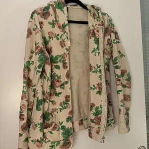 Säljer en superfin hoodie med blommigt mönster i grönt och rosa på en beige bakgrund. Den har en dragkedja framtill och är långärmad. Passar S/M