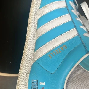 Blå och vita Adidas Gazelle sneakers - Säljer ett par snygga Adidas Gazelle sneakers i blått och vitt. Skorna har den klassiska tre-randiga designen och är perfekta för en casual look. De är i bra skick och har snörning framtill.