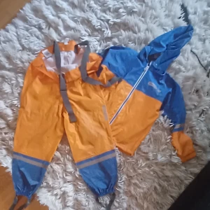 Barn. Orange och blå regnställ från Didriksons  - Använt ett fåtal gånger och är som nytt. Säljer ett färgglatt regnställ från Didriksons i orange och blått. Jackan har en praktisk huva och dragkedja framtill. Perfekt för regniga dagar och håller ditt barn torrt och bekvämt. Passar bra för utomhusaktiviteter och lek i alla väder. 🧡💙 