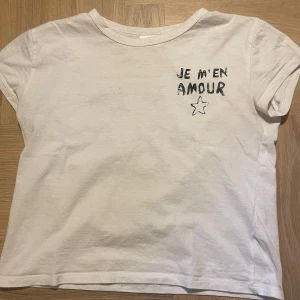 Vit t-shirt med tryck - Säljer en vit t-shirt med texten 'JE M'EN AMOUR' och en stjärna tryckt på både fram- och baksidan. Den är kortärmad och har en avslappnad passform. Perfekt för en casual look!
