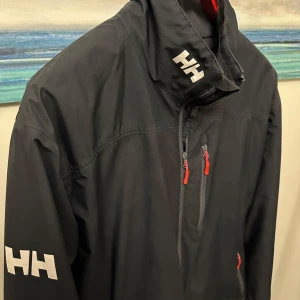 Svart vindjacka från Helly Hansen - Säljer en svart seglarjacka från Helly Hansen. Den har en hög krage med dragkedja och logotyp på både kragen och ärmen, samt på ryggen. Jackan har några år på nacken vilket gör skicket till 6/10