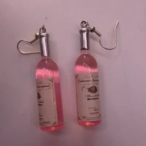Rosa vinflaska örhängen - Supercoola örhängen formade som små rosa vinflaskor med etikett. Perfekta för en festlig look eller som en rolig accessoar till vardags. De har en krok i silverfärg för enkel användning. Ett måste för vinälskare som vill sticka ut!
