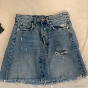 Blå jeanskjol med slitningar - Säljer en blå jeanskjol med slitningar och fransig kant. Perfekt för en avslappnad look! Kjolen har klassiska fem fickor och en knappstängning fram. Passar perfekt till sommaren eller våren. 🦋