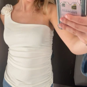 Vit oneshoulder topp med blomdetalj - Säljer en superfin vit oneshoulder topp med en söt blomdetalj på axeln. Den är gjord i ett mjukt och stretchigt material som sitter bekvämt hela dagen.